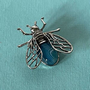 Peut inclure: Une broche en forme d'abeille argentée avec une pierre d'agate bleue et blanche au centre. Les ailes de l'abeille sont détaillées et complexes.