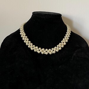 以下が含まれることがあります： マルチストランドのデザインの白い真珠のネックレス。ネックレスは小さく丸い真珠で作られており、重なり合った列のパターンで配置されています。