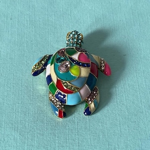 May include: A colorful turtle-shaped brooch with a mosaic design. The brooch features a variety of colors including blue, green, red, pink, and yellow. The turtle's head is adorned with small, sparkling blue gems, and the body is decorated with larger, clear gemstones.