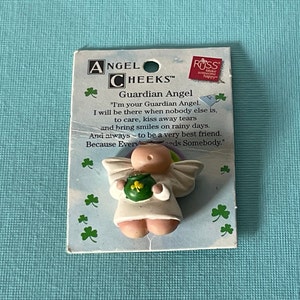 May include: A small white angel figurine with pink cheeks and a green shamrock on its chest. The angel is holding a pot of gold. The figurine is on a card that reads "Angel Cheeks Guardian Angel" and "I'm your Guardian Angel. I will be there when nobody else is, to care, kiss away tears and bring smiles on rainy days. And always to be a very best friend. Because Everyeds Somebody."