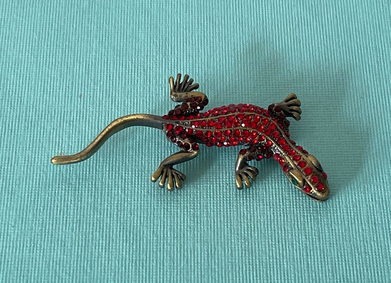 May include: A red jeweled lizard brooch with green eyes. The lizard is made of metal and has a long tail.