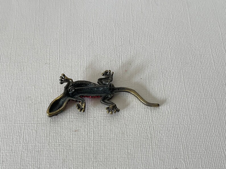 May include: A red and black jeweled gecko brooch with a metal pin on the back.