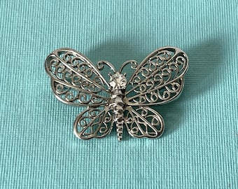 May include: A silver-toned butterfly brooch featuring intricate filigree metalwork on the wings and a single clear, round faceted rhinestone set at the center of the body. The design showcases delicate swirling patterns throughout the wings.