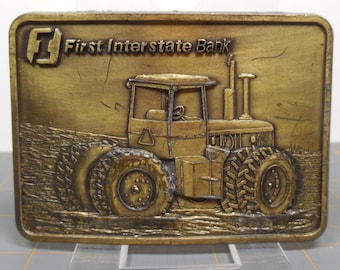 May include: A gold-toned metal belt buckle featuring a tractor with large tires and the text "First Interstate Bank".