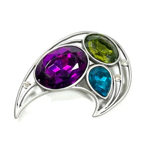 May include: Silver-toned brooch with an asymmetrical design. It features three large, faceted gemstones in purple, green, and teal, along with small clear crystals. The brooch has a curved, crescent-like shape.