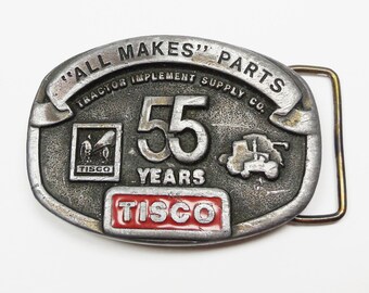 May include: A silver belt buckle with the text "All Makes Parts Tractor Implement Supply Co." and "55 Years TISCO" in red lettering. The buckle features a tractor and a logo with the text "TISCO".