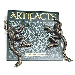 May include: A decorative plaque featuring two bronze-colored frogs climbing on a dark green background. The word "ARTIFACTS" is in gold lettering above a white splatter design. The Roman numeral "MCMLXXXVI" is at the bottom.