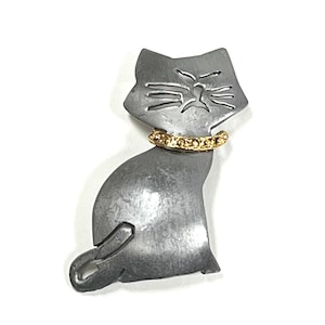 May include: A silver metal cat brooch with a gold collar. The cat is sitting with its tail curled around its body. The cat's face is detailed with cutouts for the eyes, nose, and whiskers.
