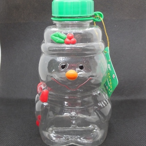 May include: Clear plastic snowman-shaped water bottle with a green lid and a red and green holly design on the hat. The bottle has a red and white candy cane design on the side. The bottle has a tag that says "Keeper's Choice".