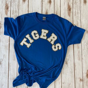 May include: A royal blue t-shirt with the word "TIGERS" in white letters with gold glitter accents.