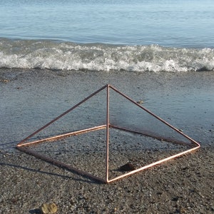 May include: A copper wire pyramid sculpture on a sandy beach with a wave in the background.