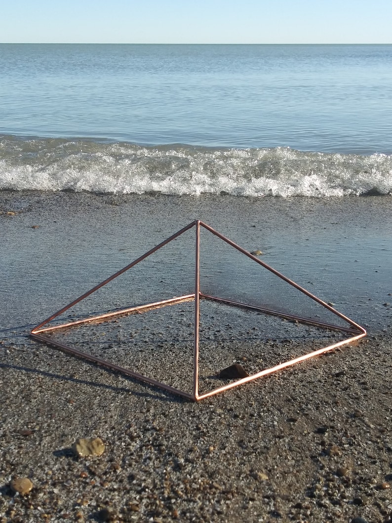 May include: A copper wire pyramid sculpture on a sandy beach with a wave in the background.