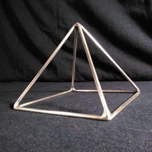 May include: A silver-colored, geometric pyramid sculpture. The pyramid is constructed from thin metal rods, forming a three-dimensional shape with a square base and four triangular sides. The sculpture is displayed against a dark background.