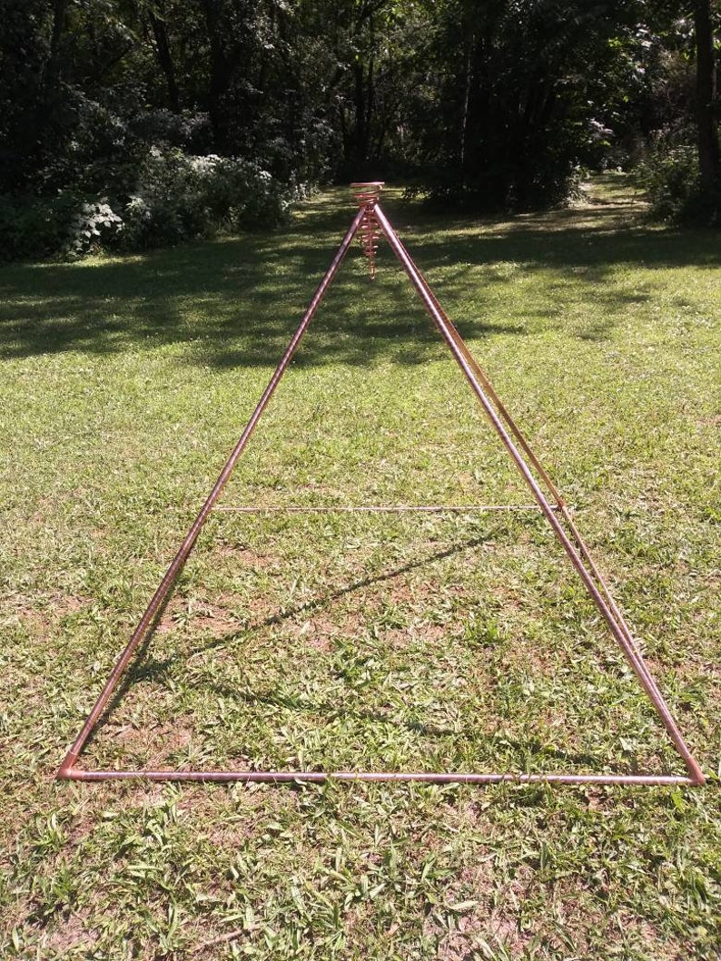 Puede incluir: Una estructura de pir&aacute;mide de cobre con un cristal colgante en el centro. La pir&aacute;mide est&aacute; de pie sobre un c&eacute;sped.