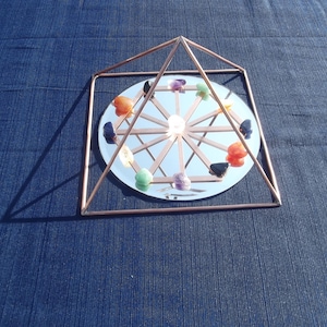 May include: A copper wire pyramid with a mirrored base and a circle of colorful gemstones arranged around the center. The pyramid is on a blue fabric surface.
