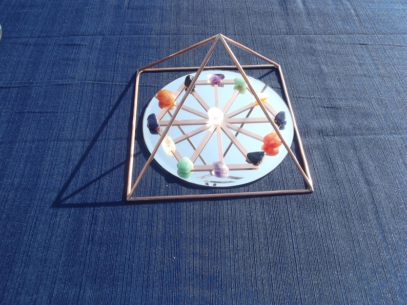 May include: A copper wire pyramid with a mirrored base and a circle of colorful gemstones arranged around the center. The pyramid is on a blue fabric surface.
