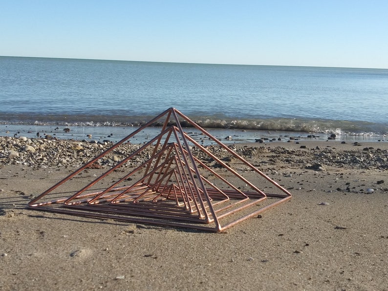 May include: A copper wire sculpture of a series of nested pyramids on a sandy beach with a blue ocean in the background.