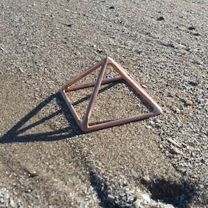 May include: A rose gold metal pyramid sits on a sandy beach. The pyramid is made of thin wire and has a geometric shape.