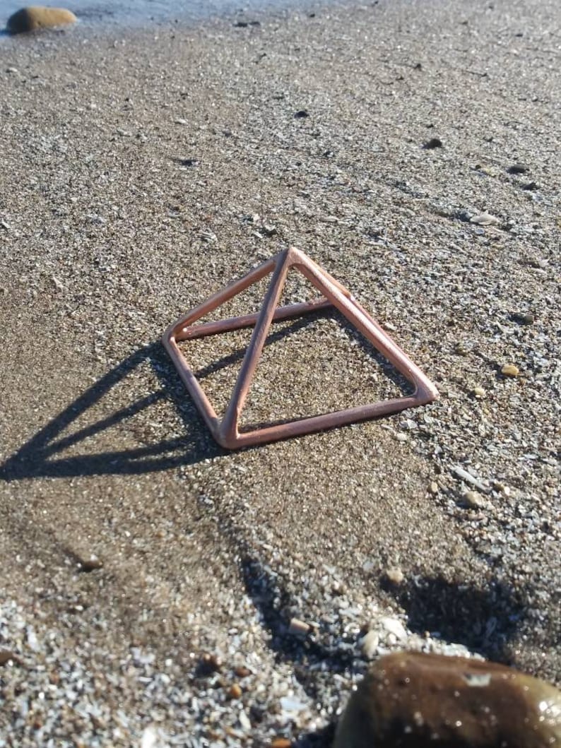 May include: A rose gold metal pyramid sits on a sandy beach. The pyramid is made of thin wire and has a geometric shape.
