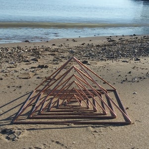May include: A copper pyramid sculpture with multiple layers, arranged in a decreasing size pattern, on a sandy beach.