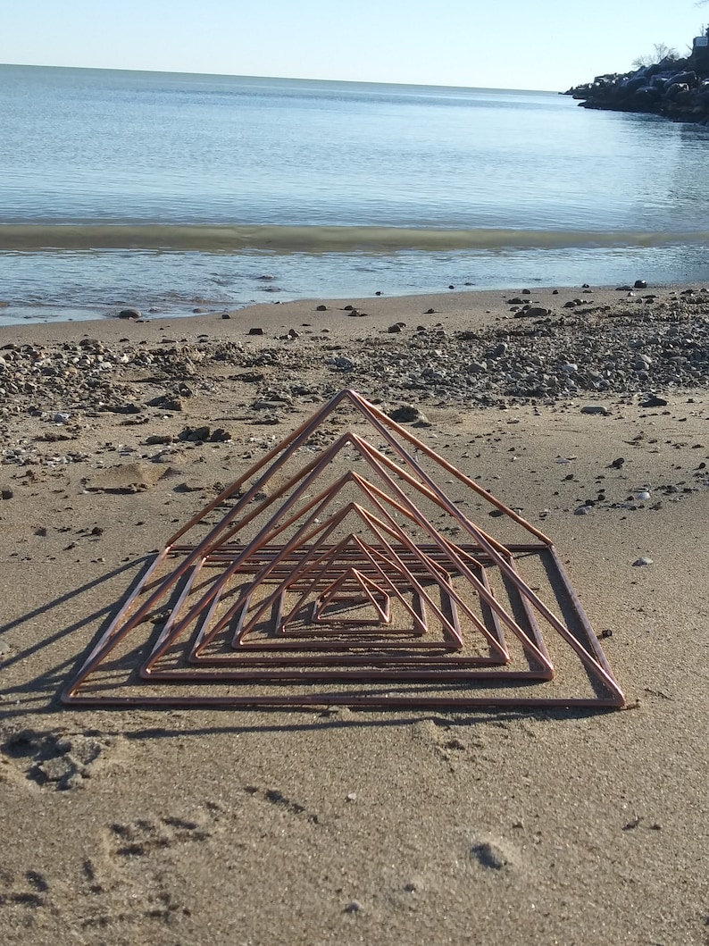 May include: A copper pyramid sculpture with multiple layers, arranged in a decreasing size pattern, on a sandy beach.