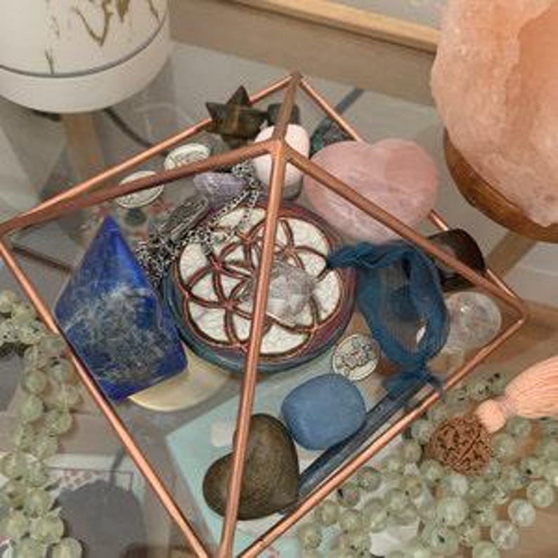 May include: A glass pyramid display case with a variety of crystals and stones, including a large rose quartz heart, a blue lapis lazuli, a clear quartz crystal, and a small blue stone. There is also a silver necklace with a pendant and a blue ribbon.