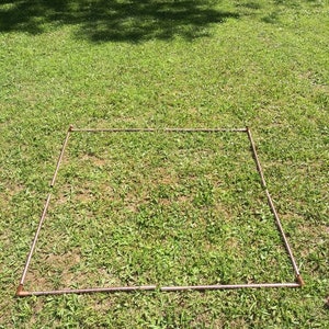 Puede incluir: Un marco cuadrado hecho de tuber&iacute;as de cobre se coloca sobre un c&eacute;sped. El marco est&aacute; hecho de cuatro tuber&iacute;as de igual longitud, cada una con una curva de 90 grados en cada extremo. El marco est&aacute; colocado plano sobre el c&eacute;sped, con las tuber&iacute;as formando una forma cuadrada.