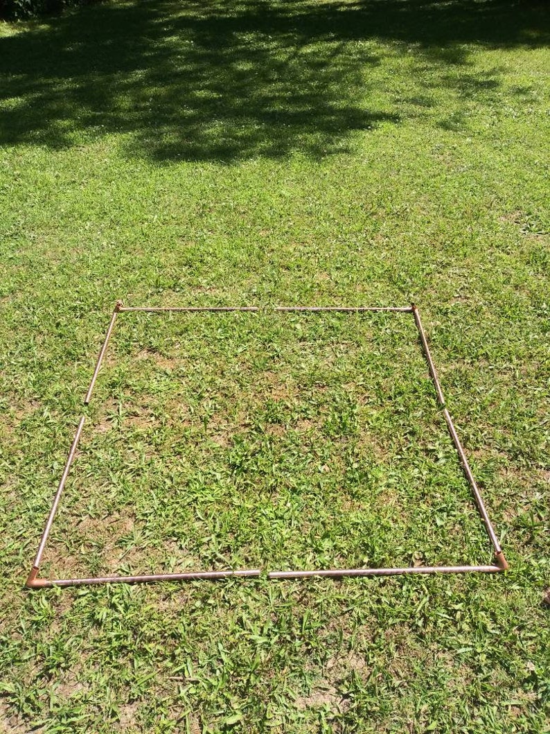 Puede incluir: Un marco cuadrado hecho de tuber&iacute;as de cobre se coloca sobre un c&eacute;sped. El marco est&aacute; hecho de cuatro tuber&iacute;as de igual longitud, cada una con una curva de 90 grados en cada extremo. El marco est&aacute; colocado plano sobre el c&eacute;sped, con las tuber&iacute;as formando una forma cuadrada.