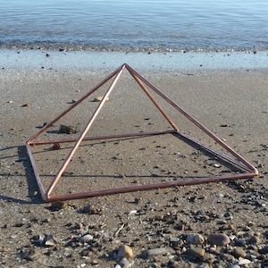 May include: A copper pyramid sits on a sandy beach with small rocks and pebbles. The pyramid is made of thin copper tubing and is open on all sides.