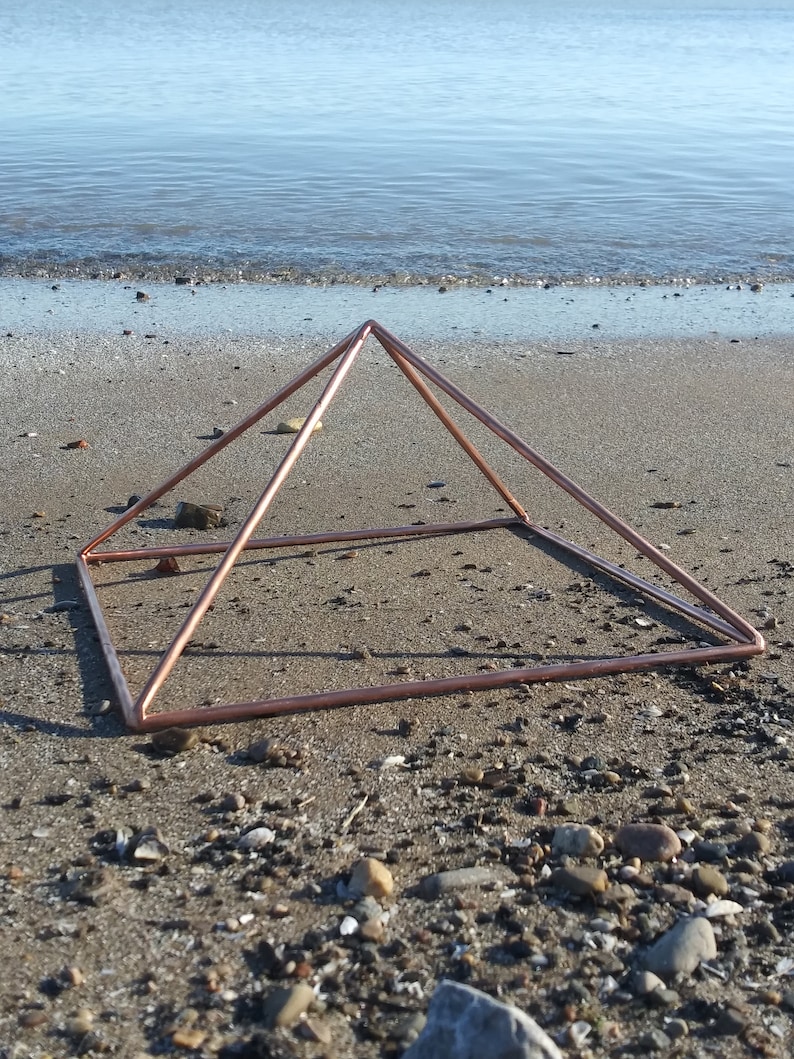 May include: A copper pyramid sits on a sandy beach with small rocks and pebbles. The pyramid is made of thin copper tubing and is open on all sides.