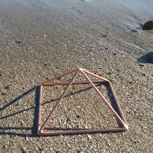 May include: A copper-colored metal pyramid frame sits on a sandy beach. The pyramid is made of thin wire and has a square base.