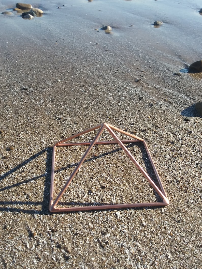 May include: A copper-colored metal pyramid frame sits on a sandy beach. The pyramid is made of thin wire and has a square base.