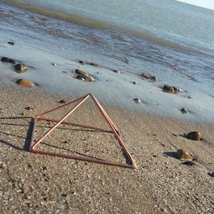 May include: A copper-colored metal pyramid sits on a sandy beach near the water's edge.