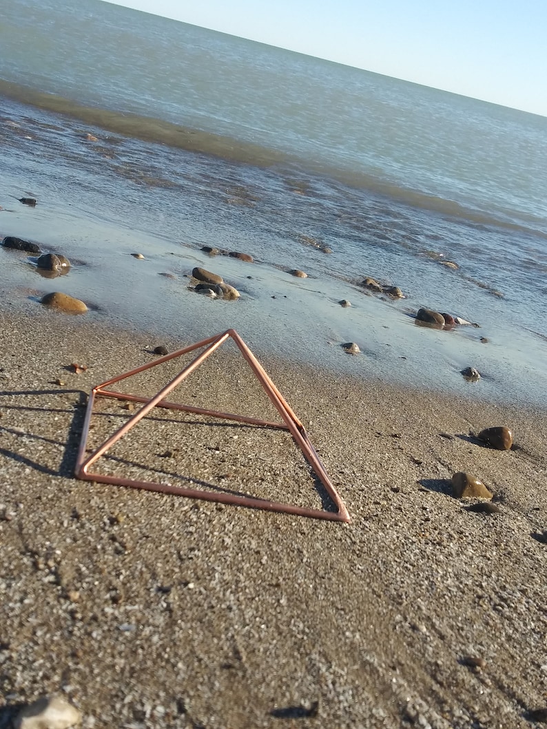 May include: A copper-colored metal pyramid sits on a sandy beach near the water's edge.