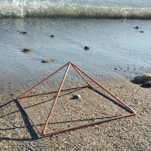 May include: A copper wire pyramid sits on a beach with small rocks and a wave in the background.