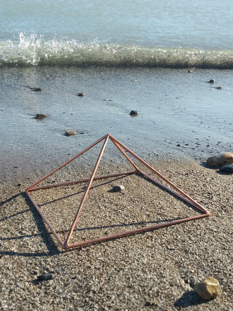 May include: A copper wire pyramid sits on a beach with small rocks and a wave in the background.