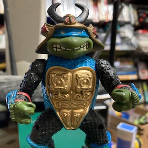May include: A Teenage Mutant Ninja Turtle action figure in a samurai-inspired outfit. The figure is primarily green, blue, and black, with gold accents on the chest. It features a horned helmet and traditional Japanese footwear.