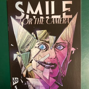 May include: A comic book titled Smile For The Camera issue number 5 with a black cover featuring a fragmented, shattered mirror effect over a person's face. The artwork uses pink, purple, and blue tones with sharp, angular shards of glass.