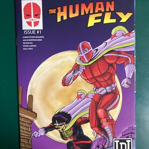 May include: Comic book cover titled "The Human Fly" featuring two costumed figures in action. The main figure is in a red suit with a white cape. The cover includes the text "Issue #1" and the names of the creators.