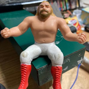 May include: A vintage action figure of a muscular wrestler with blonde hair and a beard. The figure is wearing white pants and bright red boots. The wrestler is posed with arms outstretched, sitting on a green surface.