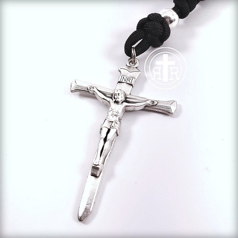 May include: Silver crucifix pendant with a depiction of Jesus Christ on the cross. The inscription "INRI" is visible on the cross. The pendant is attached to a black paracord rosary.