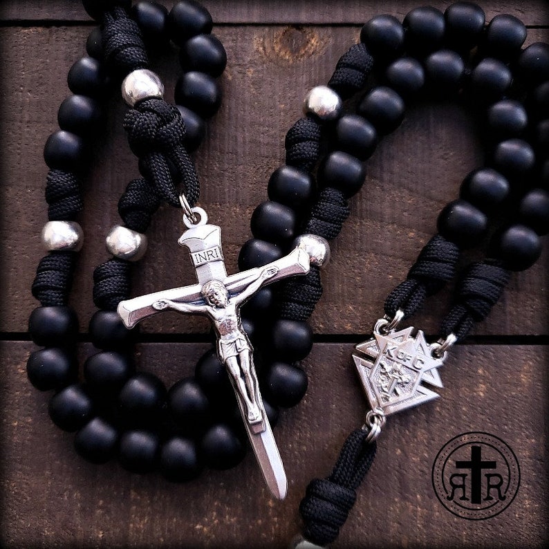 May include: Black rosary beads with a silver crucifix and a silver medal. The crucifix has the inscription "INRI" on the top. The medal has a design of a flower with the inscription "K of C" on it.