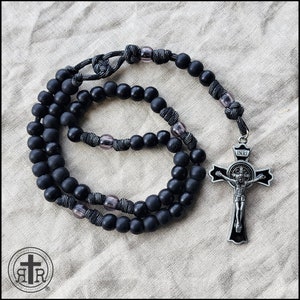 May include: Black wooden rosary beads with a silver crucifix featuring a figure of Jesus Christ. The crucifix has the inscription "INRI" above the figure. The rosary is strung with black cord and grey glass beads.
