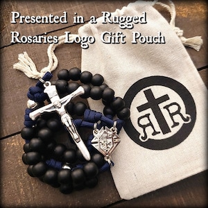 May include: Black rosary beads with a silver crucifix and a silver medal with the Knights of Columbus logo. The rosary is presented in a white drawstring bag with a black logo that says "Rugged Rosaries".