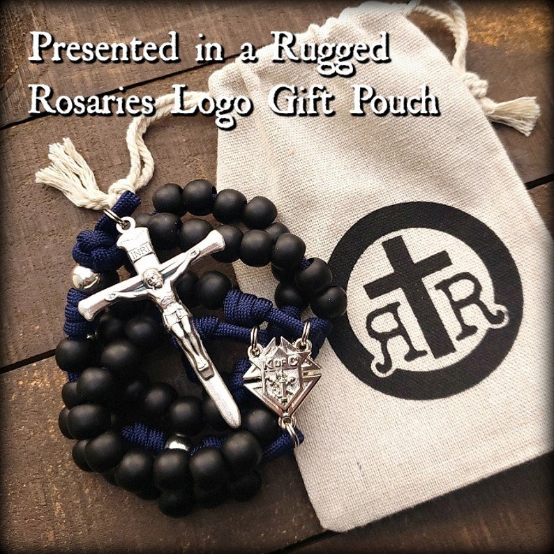 May include: Black rosary beads with a silver crucifix and a silver medal with the Knights of Columbus logo. The rosary is presented in a white drawstring bag with a black logo that says "Rugged Rosaries".