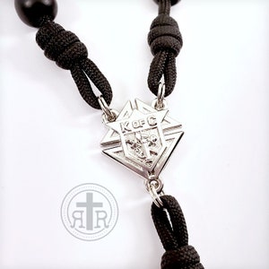 May include: Black paracord rosary with a silver pendant featuring the Knights of Columbus logo. The pendant is a diamond shape with a star at the top and the letters "K of C" in the center.