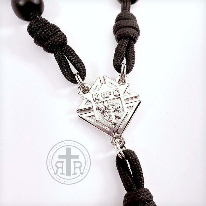 May include: Black paracord rosary with a silver pendant featuring the Knights of Columbus logo. The pendant is a diamond shape with a star at the top and the letters "K of C" in the center.
