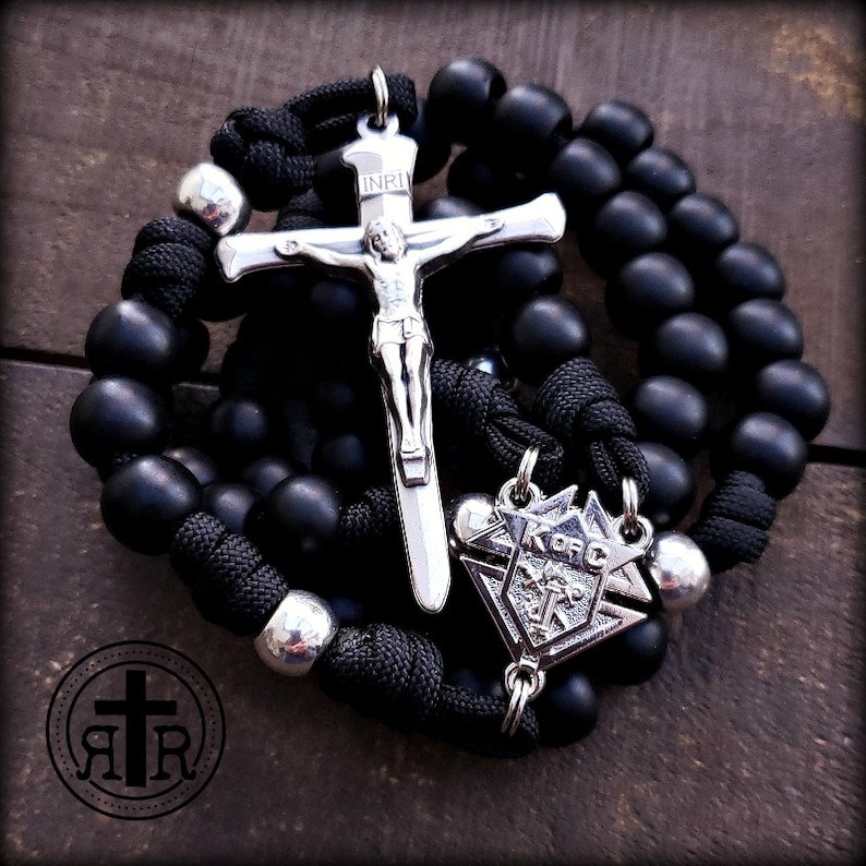 May include: Black rosary beads with a silver crucifix and a silver pendant with the letters "K of C" on it. The crucifix has the inscription "INRI" on it.