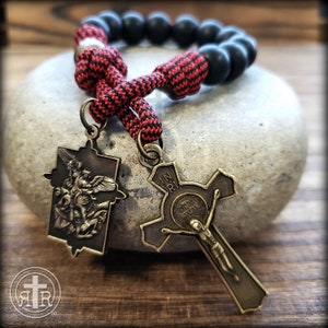 May include: A rosary with black beads and a red and black cord. The rosary features two antique-style metal pendants: a cross with a depiction of Jesus and a rectangular pendant with an angel. The rosary is displayed on a gray stone.
