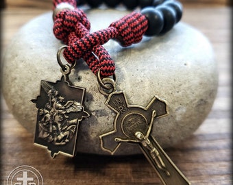 May include: A rosary with black beads and a red and black cord. The rosary features two antique-style metal pendants: a cross with a depiction of Jesus and a rectangular pendant with an angel. The rosary is displayed on a gray stone.